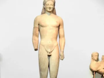 A tall marble Kouros statue of a nude male figure standing frontally with long braided hair and a missing left forearm.