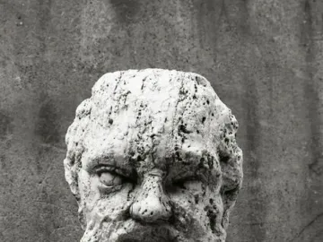 Weathered stone bust detail from the Babuino Fountain, showing a rough bearded face on a dark base.