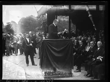 A small ornate section of the Monument to Mickiewicz is visible above a dark-draped podium as a speaker addresses a crowd.