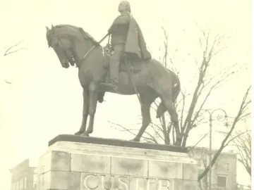 Seven uniformed men stand at attention before the Custer Monument in Monroe, Michigan, with a mounted figure atop the stone pedestal.