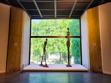 Two elongated bronze Giacometti sculptures stand on a white platform before a large window with trees outside.