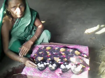 Dark padukas with toe knobs arranged on patterned cloth, with small white flowers and a woman in a green sari nearby.