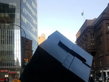 The Alamo sculpture shown as a large dark cube tilted on one corner in an urban plaza.
