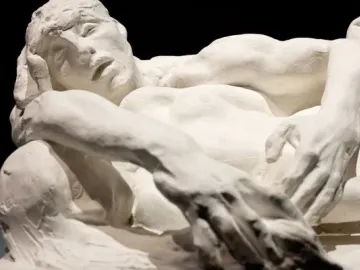 Avarice and Lust by Auguste Rodin, a cream-colored sculpture of two intertwined nude figures against a dark background.