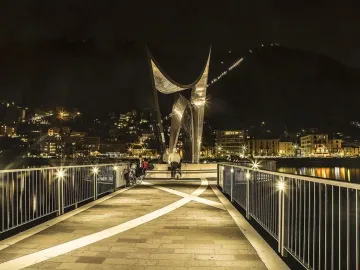 Life Electric sculpture lit at night on a waterfront promenade with Como city lights and a dark mountainside in the background.