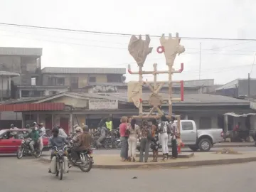 Njé Mo Yé, a tall abstract sculpture on a street median with light-colored beams, red accents, and stylized upper forms, surrounded by pedestrians and traffic.