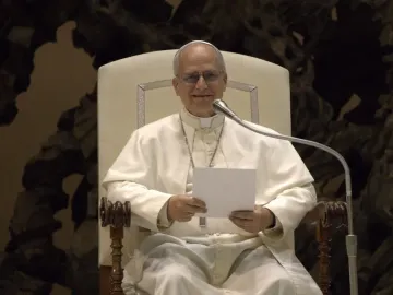A smiling pope in white clerical vestments, glasses, and a skullcap speaks into a microphone while holding a blank paper.