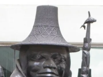 Close-up of Spirit of Haida Gwaii showing a carved figure in a woven-textured hat and patterned cloak beside a whale-topped staff.