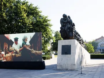 Train of Pain monument on a light stone base in a paved square, with a large historical display screen beside it.