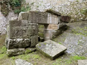 An Etruscan tomb with a small rectangular doorway in a moss-covered ashlar structure, with a fallen stone on the grass in front.