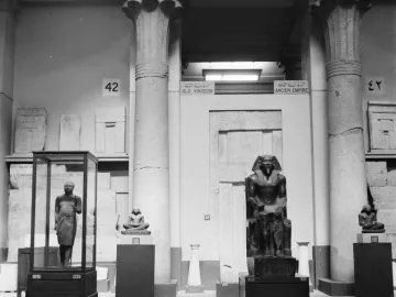 Khafre Enthroned statue displayed on a pedestal in a museum, with a falcon deity behind the pharaoh\'s head.