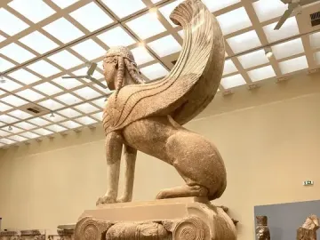 The Sphinx of Naxos shown in left profile on a fluted column inside a museum hall.