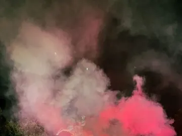 The Statue of Honor stands on a pedestal at night as red and white smoke rises around a crowd below.