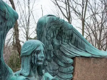 Left detail of the Haserot Angel, showing a weathered greenish winged figure holding a staff with dark streaks on its face.