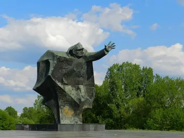 Kościuszkowców Memorial bronze figure with one arm outstretched against a blue sky and trees.