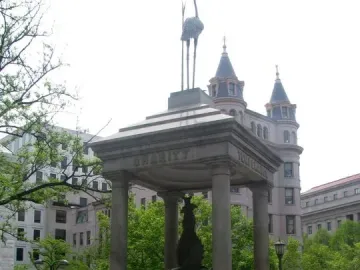 Temperance Fountain with a bird figurine on top in a paved urban plaza, with a girl standing nearby and a man walking in the background.
