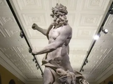 Neptune and Triton marble sculpture by Bernini shown in a museum hall, with a standing bearded figure over a kneeling figure covering his face.