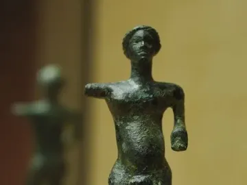 Bronze standing nude male statuette from the Neuvy-en-Sullias Hoard on a marbled base, with a similar figure blurred in the background.
