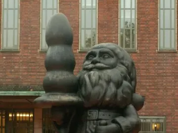 Dark Santa Claus statue holding a bell and a large black bulbous object in front of a brick building.