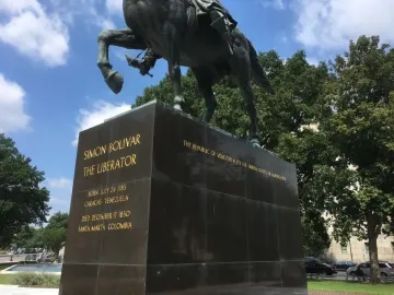 Lower section of the Simón Bolívar equestrian statue on a dark stone pedestal with gold inscriptions, framed by trees and a cloudy blue sky.