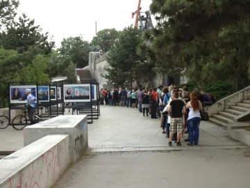 A long queue of people stands below the Prague Metronome near display panels and trees.