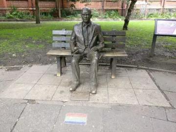 Alan Turing Memorial bronze statue seated on a bench holding an apple, with a mosaic foreground and park behind it.