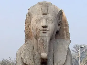 The Sphinx of Memphis, a weathered stone sphinx with a pharaonic headdress and false beard, stands on a rough base amid palm trees.
