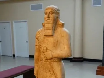 Statue of Ashurnasirpal II standing on a pedestal in a museum gallery, with a bearded figure holding an object across his chest and wearing a long patterned tunic and fringed skirt.
