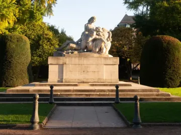 The Strasbourg War Memorial, a large stone sculpture of figures in a mournful embrace on a tiered pedestal, surrounded by grass, bushes, and autumn trees.