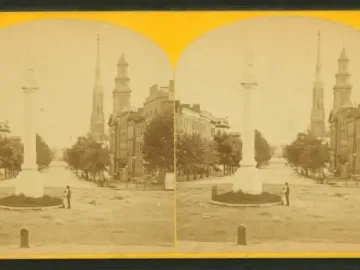 Abraham Lincoln monument on a tall column in the middle of a street, with a small figure near its fenced base and buildings in the background.
