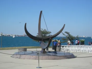 A large dark sculptural sundial stands on a circular stone base beside the water with boats in the distance.