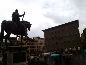 Cosimo I Equestrian Monument seen from behind in a wet Florence square with umbrellas and a Generali building in the background.