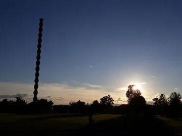 The Endless Column silhouetted against a bright evening sky with trees and grass in the foreground.