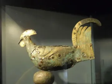 Gallo di Ramperto, a weathered metallic bird sculpture with traces of gold on its crest and tail, mounted on a tiered spherical pedestal inside a display case.