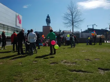 Marshal Mannerheim equestrian statue seen from behind on a tall plinth in a grassy park, with people gathered around and colorful balloons nearby.
