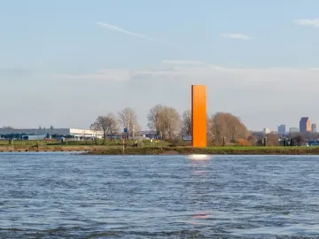 Rheinorange sculpture standing beside a wide river with sunlight reflecting on the water and distant buildings in the background.