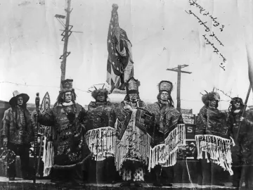 Six men in ceremonial attire stand near the Chief Seattle statue dedication site beside a flagpole.
