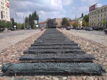Monument to the Fallen and Murdered in the East stretching along a cobblestone median in Warsaw, with dark inscribed bars leading into a jagged structure.