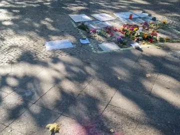 Flowers, signs, and a red candle laid on cobblestones within a green chalk outline near the Peace Statue.