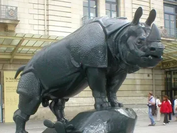 Large rhinoceros sculpture with a textured hide standing on a decorated base outdoors near a stone building.