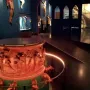 Baptismal font with reddish-brown sculpted reliefs and small animal figures around the base, displayed in a dim gallery.