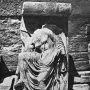 Damaged relief sculpture of Nike Untying Her Sandal, shown headless and draped in flowing robes beside a stone wall.