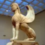 Sphinx of Naxos stone sculpture with a woman\'s head, lion\'s body, and carved wings on an ornate volute base.