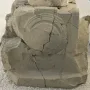 Weathered stone block with carved semicircular motifs and faint inscription lines, showing heavy cracking and damage.