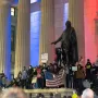 A George Washington statue stands between stone pillars as a crowd gathers below with signs and an American flag.