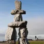 Inukshuk stone monument beside a grassy shoreline with water and distant ships under a clear blue sky.