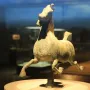Weathered bronze Flying Horse of Gansu sculpture in a galloping pose on a pedestal inside a display case.