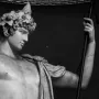 Upper body of the Antinous Braschi statue, a youthful male marble figure with a leafy wreath in his curly hair, looking down to the right and holding a staff.
