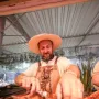 A bearded man wearing a straw hat and patterned bandana handles grilled food with tongs beside a Laçador graphic on his leather apron.