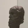 Dark patinated bronze head of the Porticello Philosopher in profile, with curly beard and hair on a light pedestal.